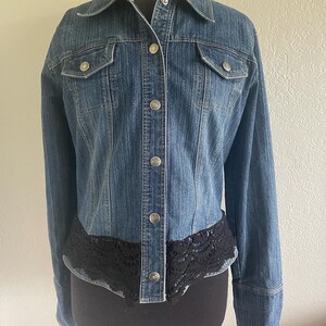 May include: A blue denim jacket with a black lace trim around the bottom. The jacket has a button-up front and long sleeves.