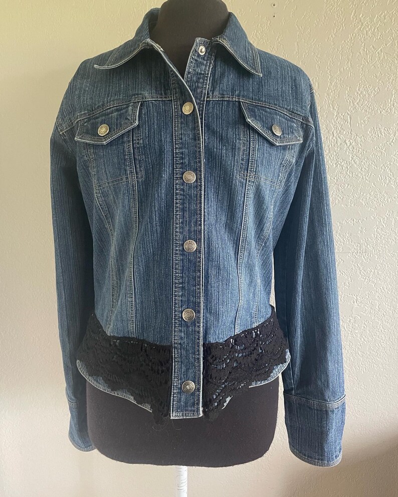 May include: A blue denim jacket with a black lace trim around the bottom. The jacket has a button-up front and long sleeves.