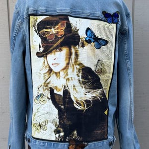 May include: A light blue denim jacket with a large image of a woman wearing a top hat with butterflies on it. The jacket has several butterfly patches on the back and sleeves.