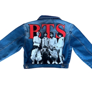 May include: A blue denim jacket with the letters "BTS" in red across the back. A group of seven people are pictured below the letters. The jacket has a classic button-up design and long sleeves.