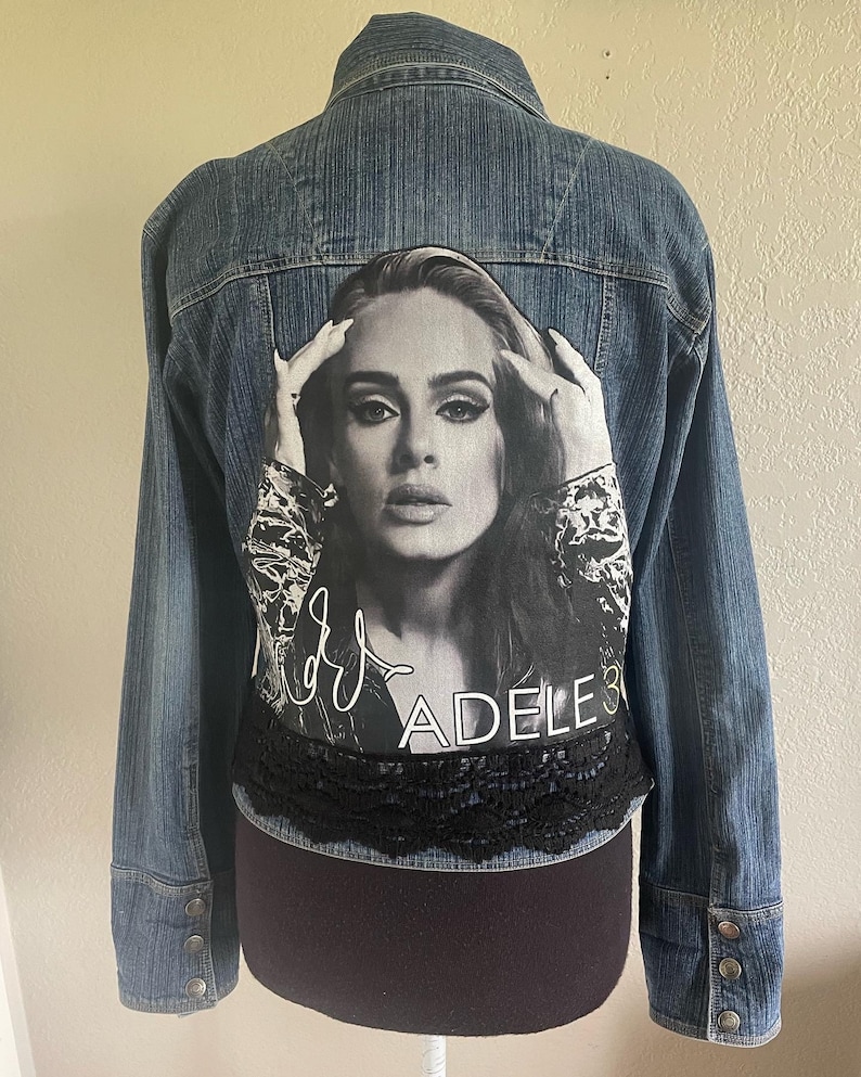 May include: A blue denim jacket with a black and white image of Adele on the back. The image includes her signature and the text "Adele 3". The jacket has a black lace trim around the bottom.