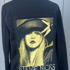 May include: Black denim jacket with a gold and black image of Stevie Nicks on the back. The image features Stevie Nicks wearing a hat and looking directly at the camera.