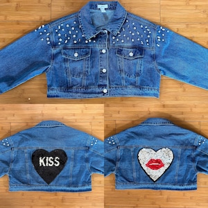 May include: A blue denim jacket with silver studs on the shoulders and a heart-shaped patch on the back. The patch is black with the word "KISS" in white letters. The jacket is cropped and has a button-up closure.