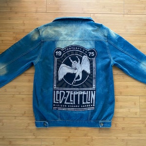 May include: A blue denim jacket with a faded wash. The back features a large graphic of the band Led Zeppelin, with the text "LED ZEPPELIN" and "MADISON SQUARE GARDEN" in white. The graphic also includes the date "FEBRUARY 7 75".