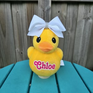 May include: A plush yellow duck toy with a white bow and the name "Chloe" in pink script. The duck has black eyes and an orange beak. The bow is adorned with small, clear embellishments.