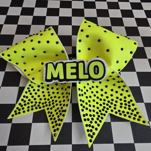 May include: A large, neon yellow hair bow with black polka dots and the name "MELO" in green letters. The bow is decorated with black rhinestones and has a white backing. The background is a black and white checkered pattern.