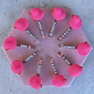 May include: A collection of pink duck-shaped hair clips arranged on a pink hexagonal surface. Each clip has a glittery tube with a different word spelled out in white beads, including "NO FEAR", "LET'S GO", and "CHEER".