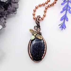 May include: A pendant necklace featuring an oval blue goldstone cabochon wrapped in copper wire. A small brass bee charm with a pale green accent sits atop the stone. The pendant hangs from a copper ball chain, showcasing intricate wire work.