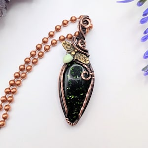 May include: A teardrop-shaped pendant featuring a dark green stone with gold flecks, encased in copper wire wrapping. The pendant includes a small brass leaf accent and a pale green bead, suspended from a copper ball chain necklace.
