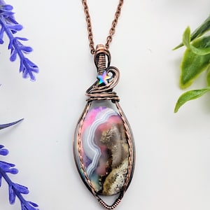 May include: A handcrafted pendant necklace featuring a large, oval-shaped stone with pink, white, and brown patterns. The stone is encased in copper wire and suspended from a copper chain. A small, iridescent star charm adds a touch of whimsy.