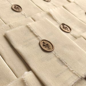 Könnte beinhalten: Off-white Stoffbeutel mit rohem Rand, versiegelt mit einem bronzefarbenen Wachssiegel. Das Siegel zeigt ein Blumendesign. Die Beutel sind in einem Raster angeordnet und erzeugen einen strukturierten, geschichteten Effekt.