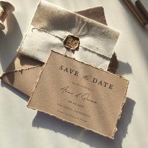 Pode incluir: Um conjunto de convites de casamento rústicos com um cartão 'Save the Date' com bordas rasgadas, um selo de cera e os nomes Anne & Quinn. O texto diz "SAVE THE DATE" e inclui a data e o local do casamento.