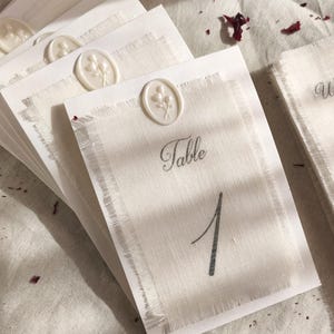 May include: A stack of ivory table cards with a linen texture, each featuring a white wax seal with a floral design. The cards are labeled with the word "Table" and a number. Scattered rose petals add a touch of color.