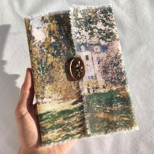 May include: A fabric-covered journal featuring a landscape painting with a bronze-colored floral clasp. The journal's edges are frayed, and the color palette includes greens, browns, and blues. The journal is held in a hand.