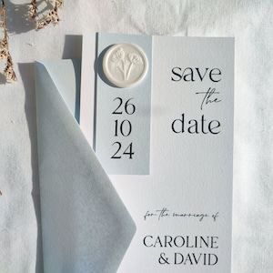 Peut inclure: Une suite de faire-part de mariage bleu clair avec une enveloppe assortie. L'invitation porte l'inscription "Save the date" et la date "26 10 24". Un sceau de cire blanc avec un motif floral est également présent.