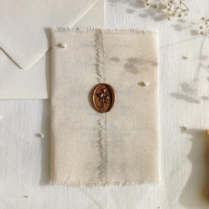 May include: A rustic wedding invitation suite featuring a cream-colored fabric with a bronze wax seal. The seal depicts a floral design. A wooden and brass wax stamp is in the lower right corner. White envelopes and floral accents complete the scene.