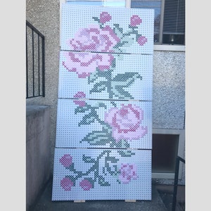 Può includere: Un grande pezzo di arte da parete a punto croce con un design floreale rosa e verde. Il design è composto da tre pannelli, ognuno con una parte diversa del design floreale. I pannelli sono montati su perni di legno.