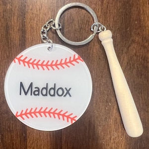 Op de afbeelding: Een honkbal-thema sleutelhanger met een witte, ronde hanger met rode stiksels en de naam "Maddox" in zwart. Een kleine houten honkbalknuppel bedel hangt naast de hanger. De sleutelhanger heeft een zilveren sleutelring en ketting.