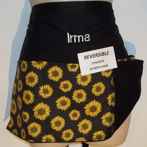May include: Reversible black apron with a sunflower pattern on the lower half. The apron has three pockets on both sides and the name "Irma" embroidered in white. The apron is displayed on a mannequin.
