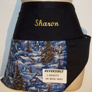 May include: Black and blue reversible apron with a winter village print on one side and a black side with the name "Sharon" embroidered in gold thread. The apron has three pockets on each side and the text "REVERSIBLE 3 POCKETS ON BOTH SIDES" printed on a white tag.