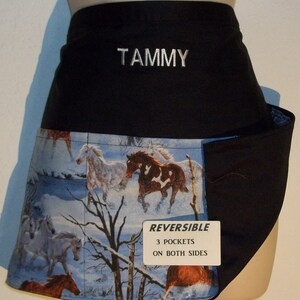 May include: Reversible black apron with a horse-themed pattern. The apron features a black top with the name "TAMMY" embroidered in white. The bottom half displays a winter scene with running horses. The apron has three pockets on both sides.