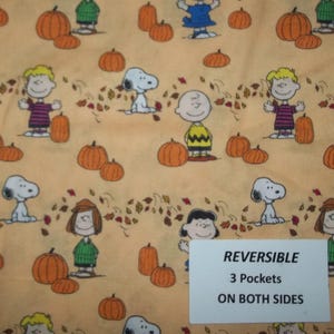 May include: Fabric with a repeating pattern of orange pumpkins, Snoopy, Charlie Brown, and other Peanuts characters on a light orange background. The fabric has the text "REVERSIBLE 3 Pockets ON BOTH SIDES" on a white rectangle.