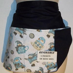 May include: A reversible black and white apron with a snowman and hot cocoa print. The apron has three pockets on both sides. The text "REVERSIBLE 3 POCKETS ON BOTH SIDES" is printed on a white tag.