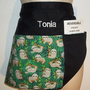 May include: Black and green reversible apron with a sloth print on one side and a black pocket on the other. The apron has a black waistband with the name "Tonia" embroidered on it. The apron has a tag that says "REVERSIBLE 3 POCKETS ON BOTH SIDES".