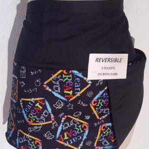 May include: A reversible black apron with three pockets. The apron features a colorful pattern with pencils, the words "Learn" and "Create", and simple math equations. A tag indicates the apron is reversible.