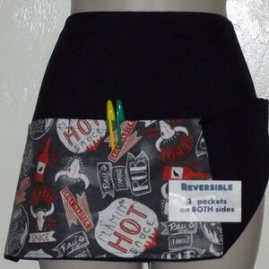 May include: Black reversible apron with a large pocket on the front. The pocket has a black fabric flap and is lined with a colorful fabric featuring a grill master theme with red, white, and black text. The text includes "Hot Sauce", "Grill Master", "Ray's Famous Rub", and "Flaming Hot Sauce". The apron has the text "REVERSIBLE 3 pockets on BOTH sides" printed on a white tag.