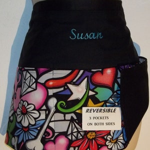 May include: Black reversible apron with a colorful graphic print of hearts, stars, and flowers. The apron has three pockets on both sides and is personalized with the name "Susan".