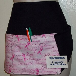 May include: A black and pink reversible apron with three pockets on both sides. The pink side features a pink ribbon pattern with the words "Love", "Hope", "Strength", "Courage", "Survive", "Cure", "Overcome", and "Believe". The apron has a label that says "REVERSIBLE 3 pockets on BOTH sides".