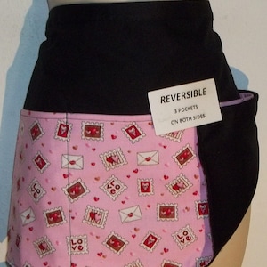 May include: A reversible black and pink apron with a pink and white print of hearts and envelopes. The apron has three pockets on both sides. The text "REVERSIBLE 3 POCKETS ON BOTH SIDES" is printed on a white tag.