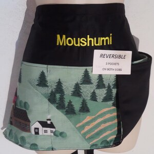 May include: A black waist apron featuring the embroidered yellow name Moushumi and a light green panel with a printed landscape of pine trees and houses. A white tag attached to the front reads REVERSIBLE 3 POCKETS ON BOTH SIDES.