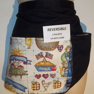 May include: A black reversible apron with three pockets on both sides. The apron features a white fabric with a colorful print of chickens, apples, hearts, and American flags. The text "REVERSIBLE", "3 POCKETS", and "ON BOTH SIDES" is printed on a white tag attached to the apron.