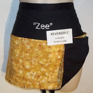May include: Reversible black and gold apron with star pattern and the name "Zee" embroidered in white. Features three pockets on both sides. The apron is designed to be worn around the waist.