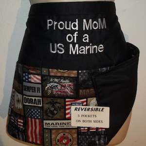 May include: Black and camouflage reversible apron with the text "Proud Mom of a US Marine" embroidered on the front. The back of the apron features a patchwork design of American flags, eagles, and military insignia. The apron has three pockets and the text "REVERSIBLE 3 POCKETS ON BOTH SIDES" printed on a white tag.