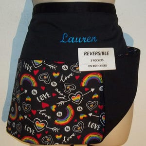 May include: Black and rainbow reversible apron with three pockets on each side. The apron has a rainbow and heart print with the text "Love is Love" and the name "Lauren" embroidered on the front.