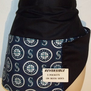 May include: A reversible black and blue apron with a Seattle Mariners logo print. The apron has three pockets on both sides. The text "REVERSIBLE", "3 POCKETS", and "ON BOTH SIDES" is printed on a white tag.