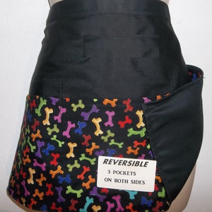 May include: Black reversible apron with three pockets on both sides. One side features a black fabric, the other side has a black background with colorful bone-shaped patterns. The apron has a label that reads "REVERSIBLE 3 POCKETS ON BOTH SIDES".
