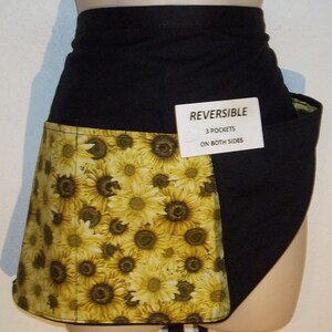 May include: Black and yellow reversible apron with a sunflower print on one side and solid black fabric on the other. The apron has three pockets on both sides. The text "REVERSIBLE 3 POCKETS ON BOTH SIDES" is printed on a white tag.