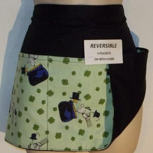 May include: A reversible black and green apron with a clover pattern and a cartoon character design. The apron has three pockets on both sides. The tag reads "REVERSIBLE 3 POCKETS ON BOTH SIDES".