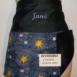 May include: Reversible apron with a black waistband and pockets. The lower portion features a colorful celestial pattern with yellow stars and blue and purple accents. The name "Jami" is embroidered in cursive. The apron has three pockets on both sides.