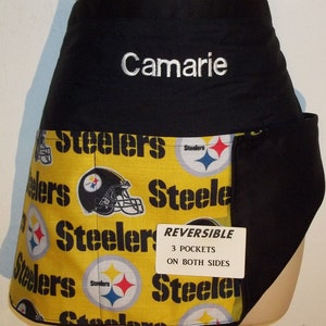 May include: Black and yellow reversible apron with the Pittsburgh Steelers logo and the word "Steelers" repeated on the fabric. The apron has three pockets on both sides and is personalized with the name "Camarie".