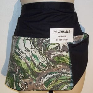 May include: Black and green reversible apron with three pockets on both sides. The apron has a marbled pattern on one side and a solid black fabric on the other side. The apron is made of a durable, water-resistant material.