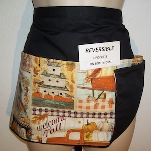 May include: A black reversible apron with a fall-themed print featuring pumpkins, birds, and a welcome fall sign. The apron has three pockets on each side, as indicated by the label "REVERSIBLE 3 POCKETS ON BOTH SIDES".