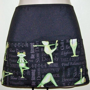 May include: Black apron with a pocket and a colorful print of frogs doing yoga poses. The print includes the words "stretch", "slow down", "breathe deeply", "find balance", and "lighten up".