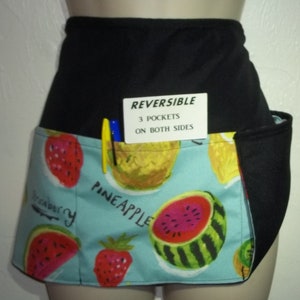 May include: A black and blue reversible apron with three pockets on both sides. The front of the apron features a blue background with a pattern of watermelons, strawberries, pineapples, and kiwi. The text "Pineapple" and "Strawberry" are visible on the apron.