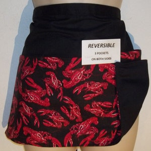 May include: Black and red reversible apron with a lobster print. The apron has three pockets on both sides. The text "REVERSIBLE" and "3 POCKETS ON BOTH SIDES" is printed on a white tag.