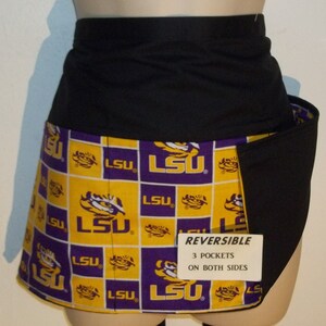 May include: A reversible black and yellow apron with a purple and yellow LSU logo pattern. The apron has three pockets on each side and the text "REVERSIBLE 3 POCKETS ON BOTH SIDES" printed on a white tag.